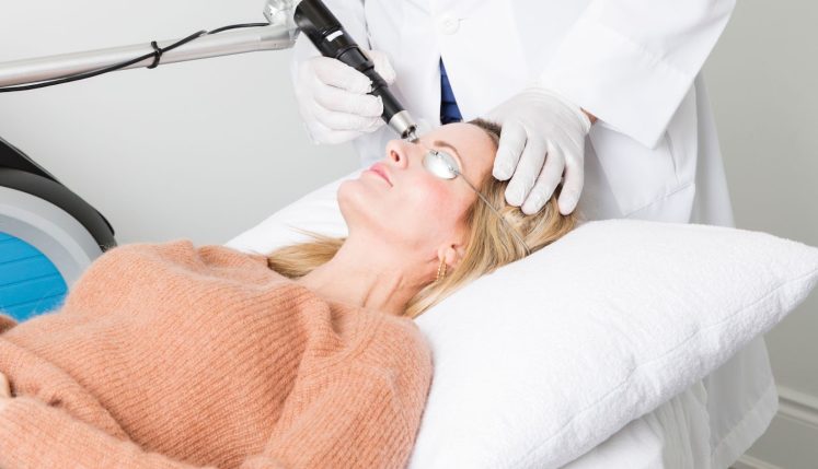 A woman receiving a laser treatment while lying on a treatment table, wearing protective eyewear, with a technician operating the laser device.