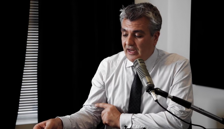 Man in a striped shirt and tie speaking into a microphone while pointing at a document during a discussion.