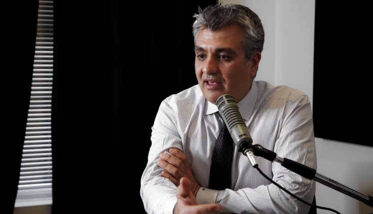 Man in a striped shirt speaking into a microphone during a discussion, with a serious expression and arms crossed.